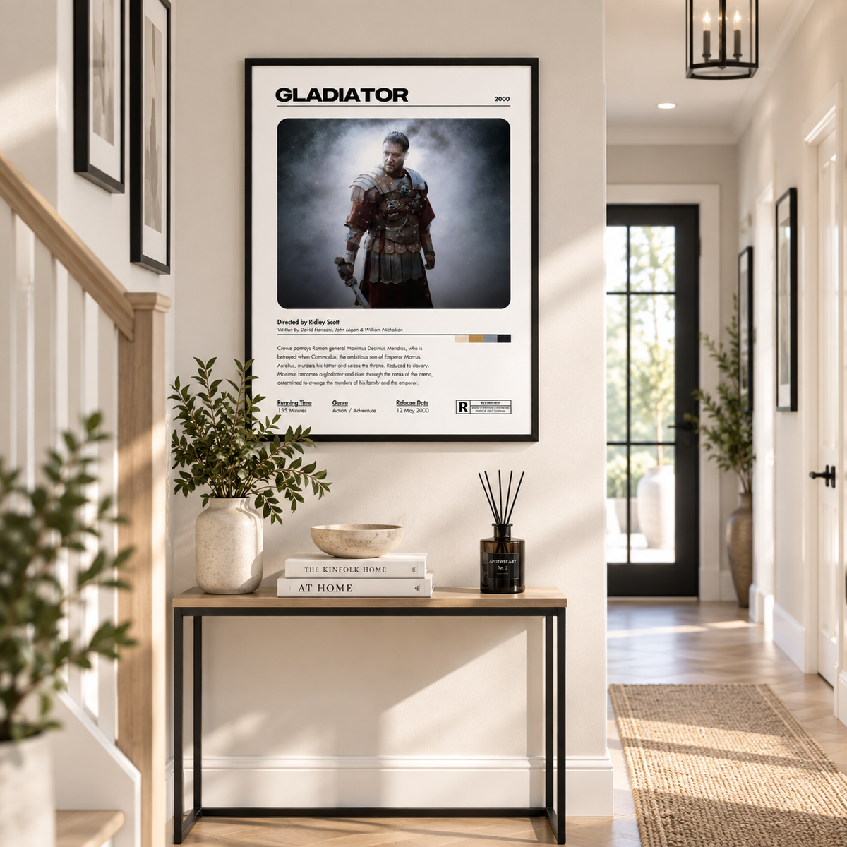 A framed Gladiator movie poster hangs on a white hallway wall above a console table with books, a vase, and a reed diffuser. The poster features a portrait of a gladiator in armor with the title 'GLADIATOR' at the top and release details below.