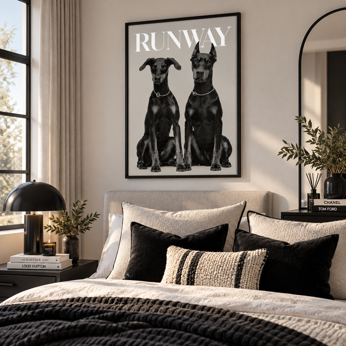 A framed black-and-white poster featuring two dogs with the word "RUNWAY" above them, displayed above a bed in a styled bedroom. The room includes a beige upholstered headboard, layered black and cream pillows, a dark lamp, books, and plants.
