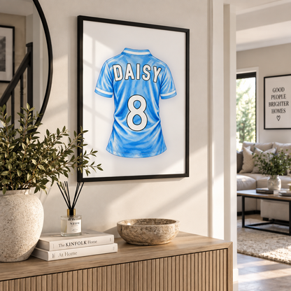 A framed custom jersey print featuring a light blue soccer-style shirt with the name DAISY and number 8 on the back, displayed on a wall in a modern living room setting above a wooden console table.