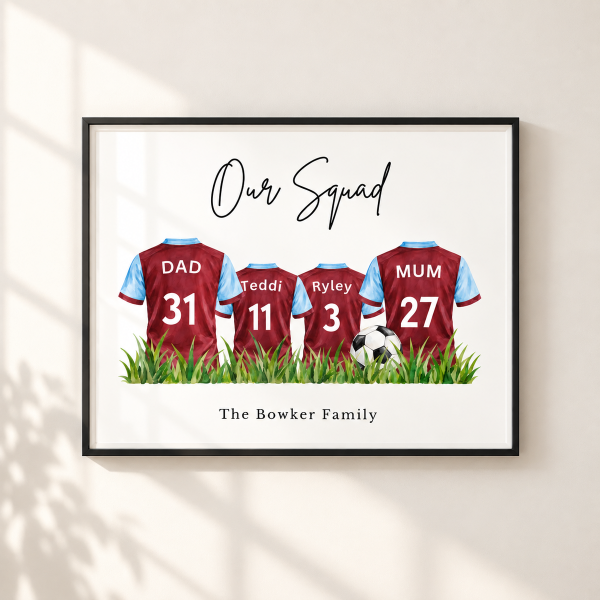 A framed personalized family soccer print on a light beige wall with soft window shadows. The print features four maroon and light blue football jerseys labeled DAD 31, Teddi 11, Ryley 3, and MUM 27, standing in green grass under the text 'Our Squad' and 'The Bowker Family'.