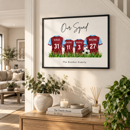 A framed personalized family print hanging on a wall in a bright living room. The artwork shows three maroon soccer jerseys with names and numbers: Dad 31, Reddi 11, and Ryley 27, with the text 'Our Squad' at the top and 'The Bowker Family' at the bottom.