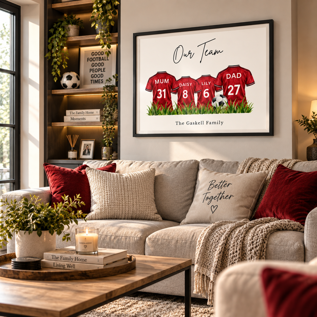 A framed personalized football jersey print hangs on a wall above a beige sofa in a styled living room. The artwork shows three red jerseys with the names Mum, Dads, and Lily, numbers 31, 8, and 27, on a white background with the text 'Our Team' and 'The Gaskell Family'.