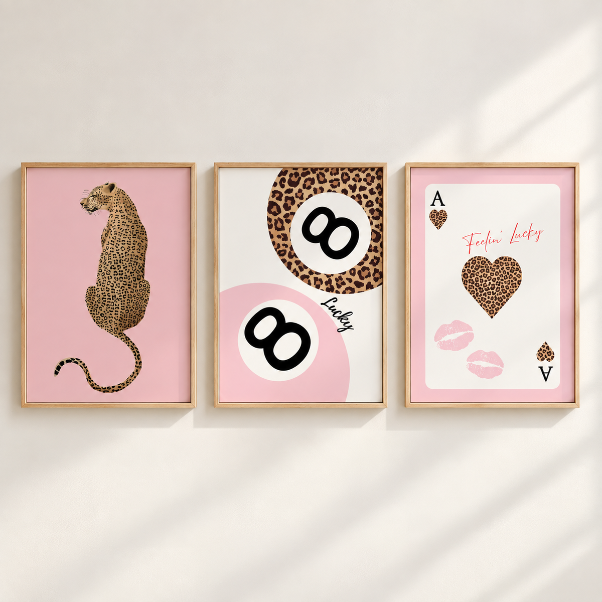 Three framed art prints with a cohesive pink and leopard-print theme, displayed side-by-side on a light beige wall with soft diagonal shadows. The left print shows a leopard in profile on a pink background; the middle features a leopard-patterned '8' on a white and pink circle with the word 'Lucky'; the right print is a pink 'Ace of Hearts' card design with a leopard heart and 'Fierce Lucky' text.