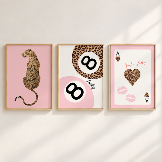 Three framed art prints with a cohesive pink and leopard-print theme, displayed side-by-side on a light beige wall with soft diagonal shadows. The left print shows a leopard in profile on a pink background; the middle features a leopard-patterned '8' on a white and pink circle with the word 'Lucky'; the right print is a pink 'Ace of Hearts' card design with a leopard heart and 'Fierce Lucky' text.