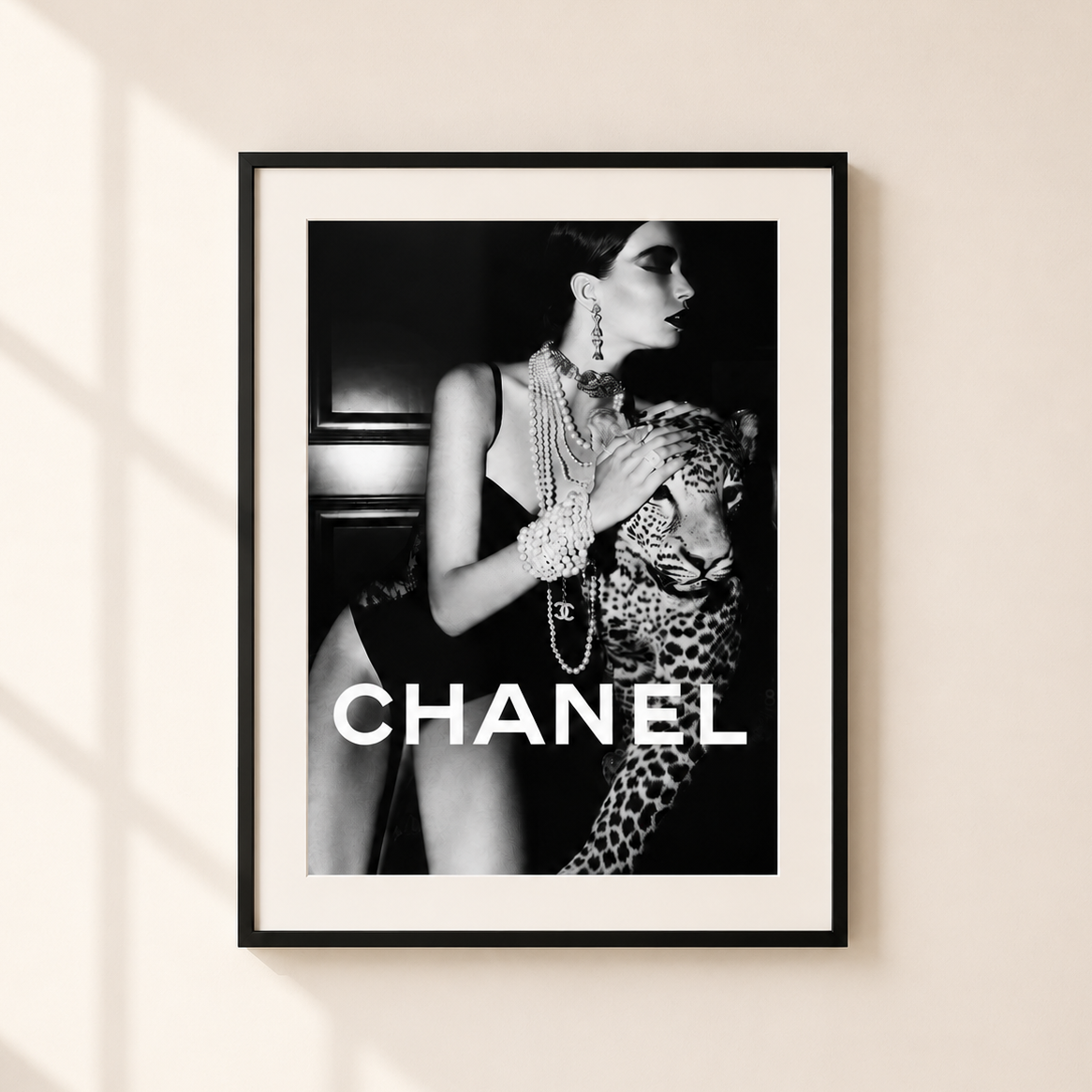 A framed black and white fashion poster featuring a woman with a leopard and the word CHANEL in large white letters, hung on a light beige wall with soft window shadows.
