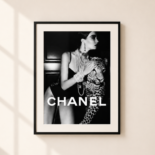 A framed black and white fashion poster featuring a woman with a leopard and the word CHANEL in large white letters, hung on a light beige wall with soft window shadows.
