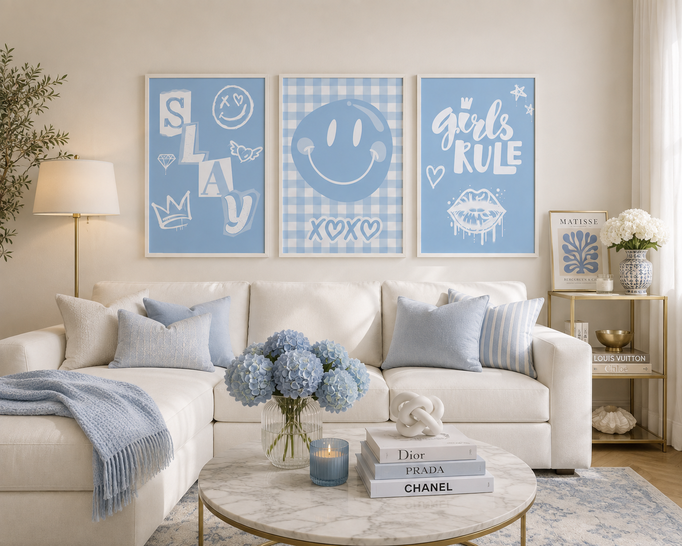 Three framed blue wall art prints hang above a white sectional sofa in a bright living room. The prints feature white graphic designs reading 'SLAY', a smiley face, and 'girls RULE'.