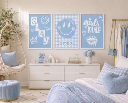 Three framed light blue wall art prints hang above a white dresser in a styled bedroom. The prints feature white graphic designs: 'SLAY' with a smiley face, a blue gingham smiley face with 'XOXO', and 'girls RULE' with lips.