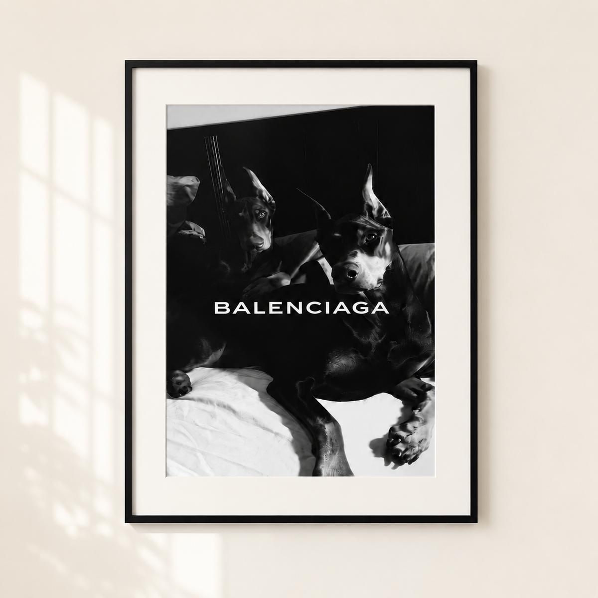 A framed black and white Balenciaga poster featuring a dramatic photo of two dogs with sunglasses, displayed on a light beige wall with soft window shadow overlays.
