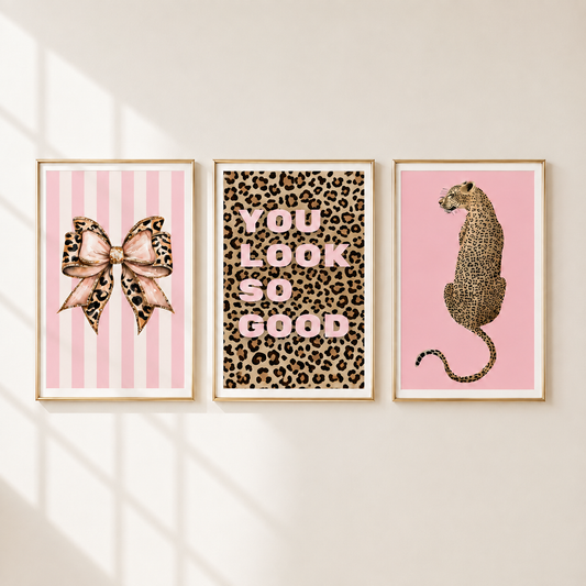 Three framed wall art prints hang on a light beige wall with soft window-shadow lighting. The left print shows a pink striped bow with leopard print details; the center print reads 'YOU LOOK SO GOOD' in pink letters over a leopard pattern; the right print shows a pink leopard illustration.