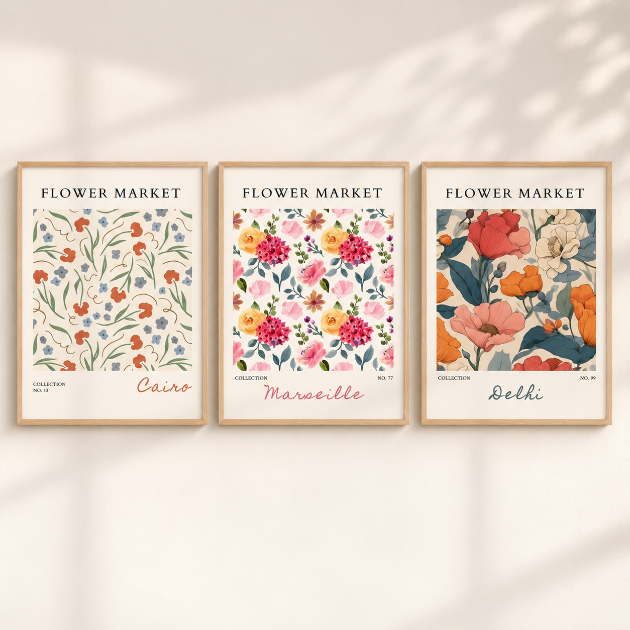 Three framed floral art prints hang side by side on a light beige wall with soft leaf shadows. Each print has a light wood frame and the header 'FLOWER MARKET' above colorful botanical illustrations and city names: Cairo, Marseille, and Delhi.