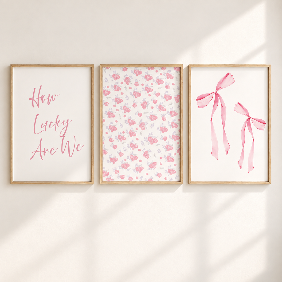 Three framed wall art prints hanging side by side on a warm off-white wall, with soft diagonal sunlight casting shadows. The left print has pink script text reading 'How Lucky Are We' on a white background.