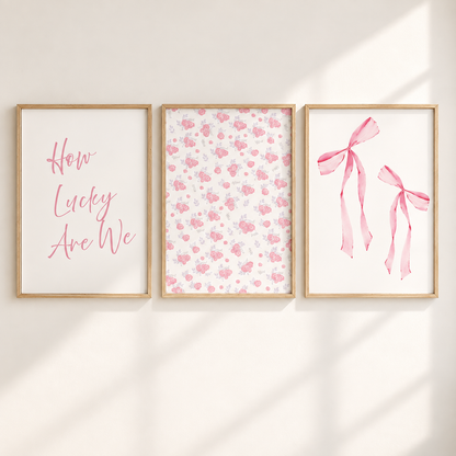 Three framed wall art prints hanging side by side on a warm off-white wall, with soft diagonal sunlight casting shadows. The left print has pink script text reading 'How Lucky Are We' on a white background.