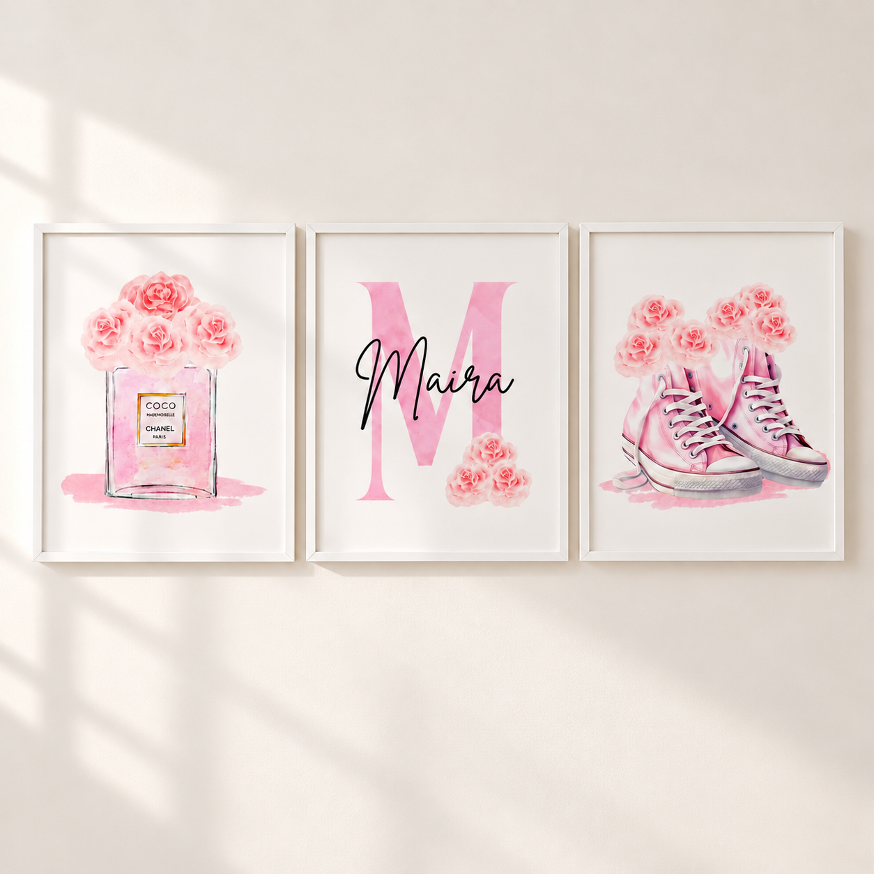 Three framed watercolor-style wall art prints hang on a light beige wall. The left print shows pink roses in a perfume bottle labeled Chanel; the center print features a large pink letter M with the name 'Maira' in black script; the right print shows pink high-top sneakers with pink roses.