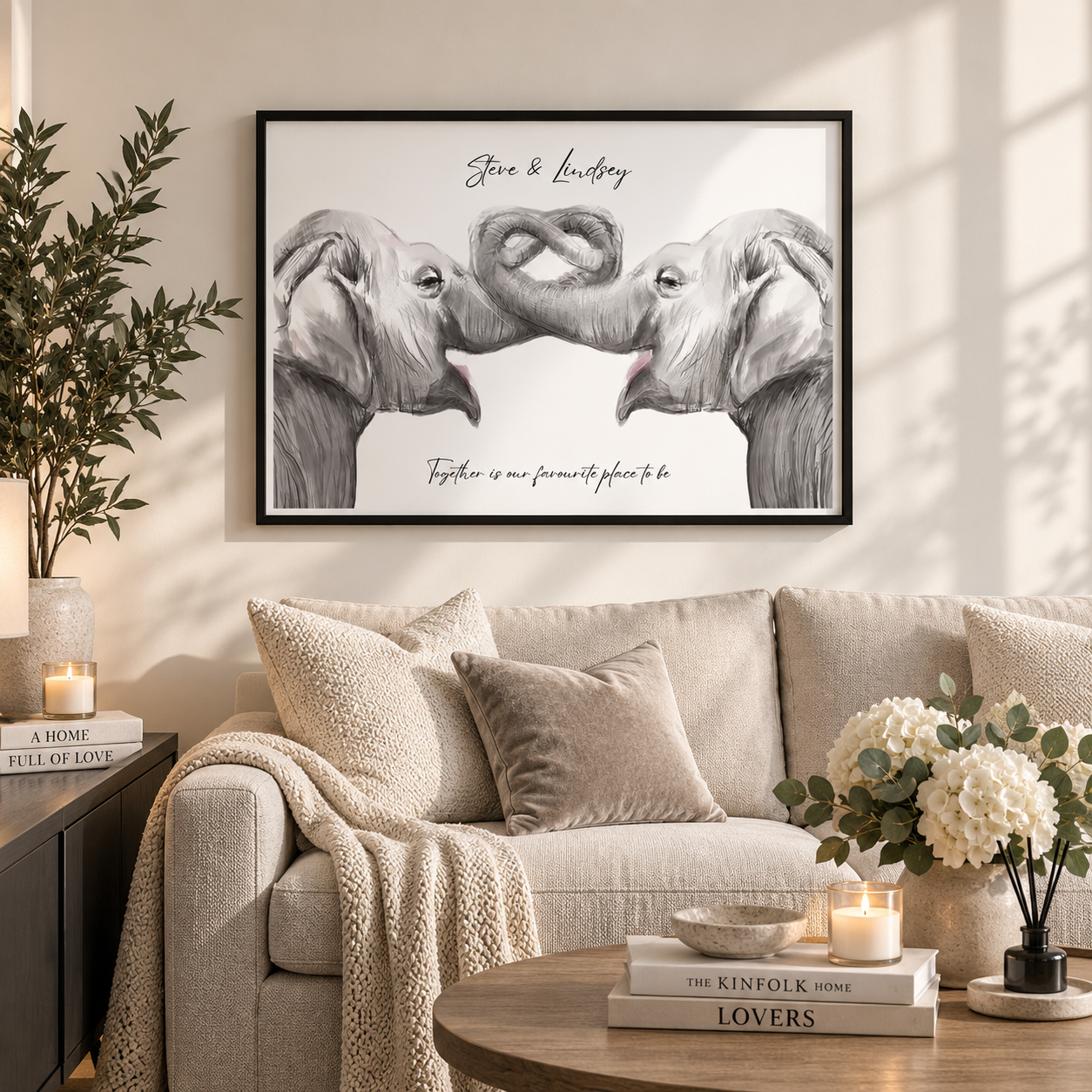 A framed black-and-white elephant artwork hangs above a beige sofa in a styled living room. The print shows two elephants with intertwined trunks and the text 'Steve & Lindsay' and 'Together is our favorite place to be.