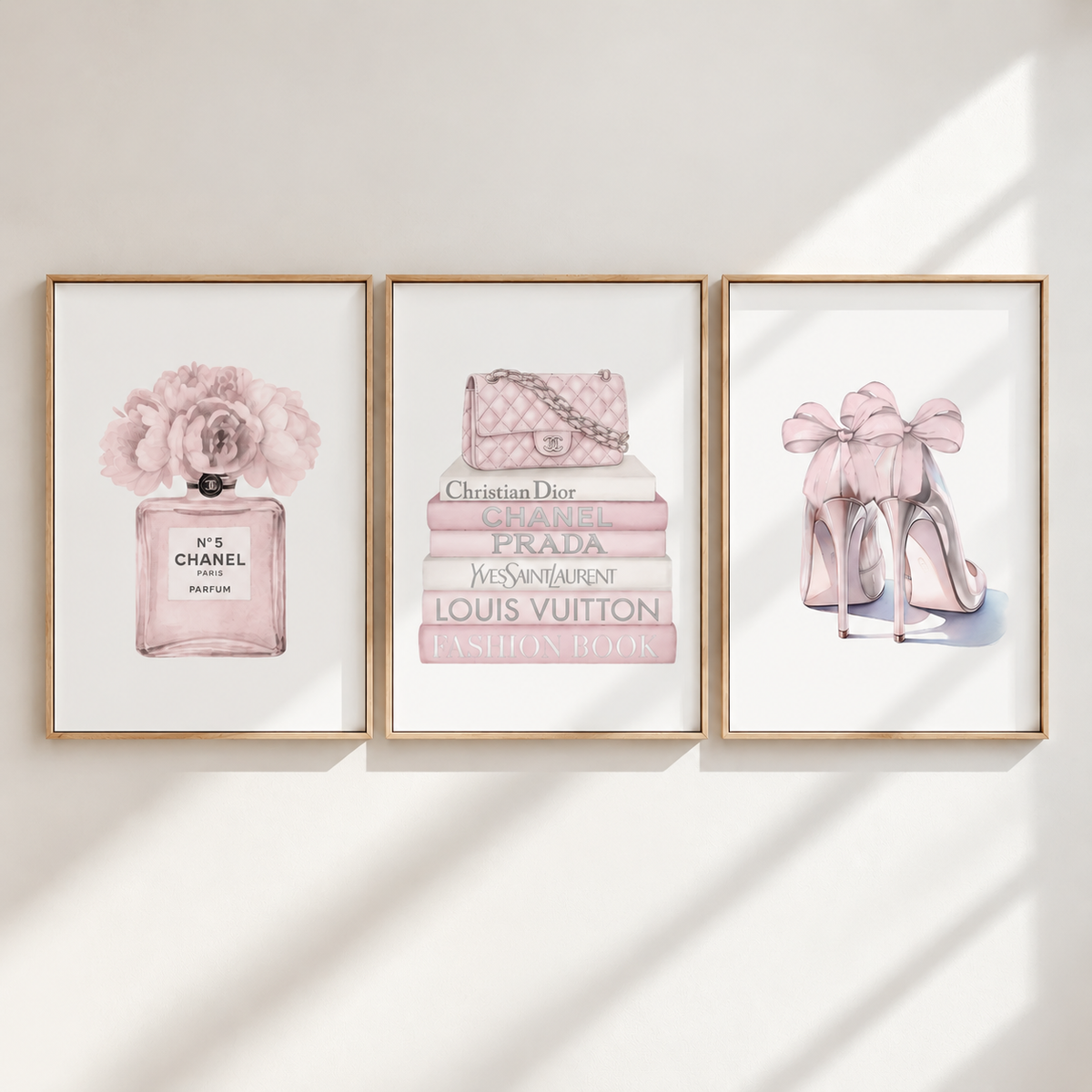 Three framed wall art prints hang on a light wall with soft diagonal shadows. The prints feature pink fashion illustrations: a Chanel perfume bottle with flowers on the left, a stack of designer books and a quilted handbag in the center, and pink high heels with large bows on the right.