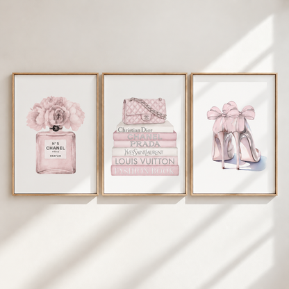 Three framed wall art prints hang on a light wall with soft diagonal shadows. The prints feature pink fashion illustrations: a Chanel perfume bottle with flowers on the left, a stack of designer books and a quilted handbag in the center, and pink high heels with large bows on the right.
