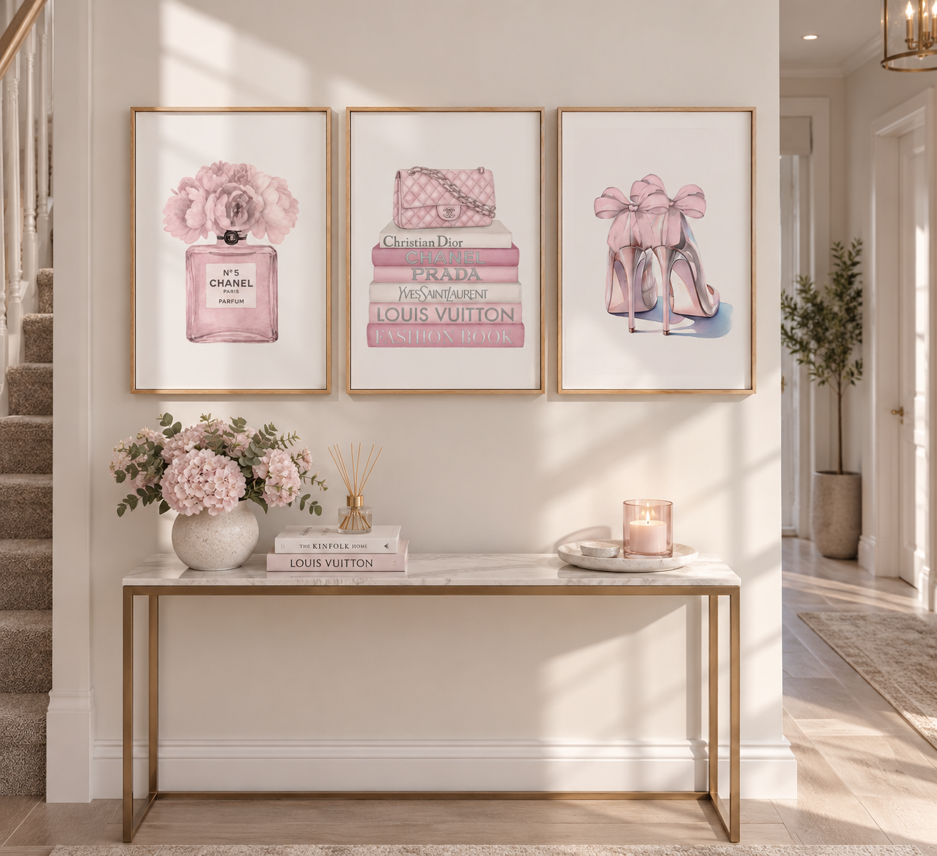 Three framed art prints with pink and gold Chanel-themed illustrations hang on a cream wall above a console table decorated with flowers and books.