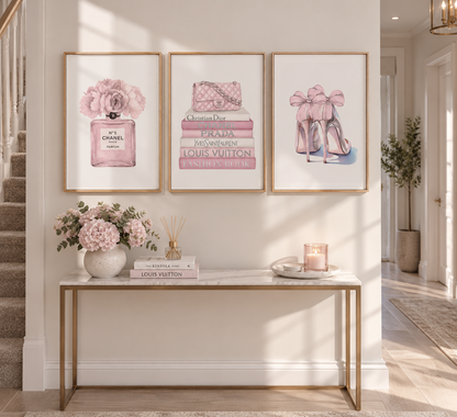 Three framed art prints with pink and gold Chanel-themed illustrations hang on a cream wall above a console table decorated with flowers and books.