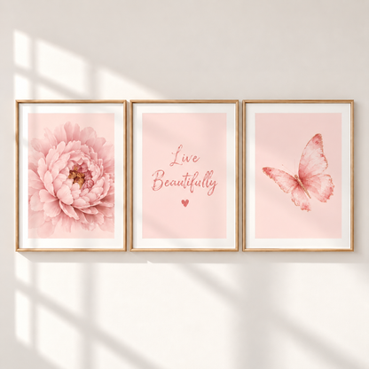 Three framed wall art prints in a row on a white wall, lit by soft window shadows. The left print shows a large pink flower, the center print reads 'Live Beautifully' in pink script with a small heart, and the right print shows a pink butterfly.