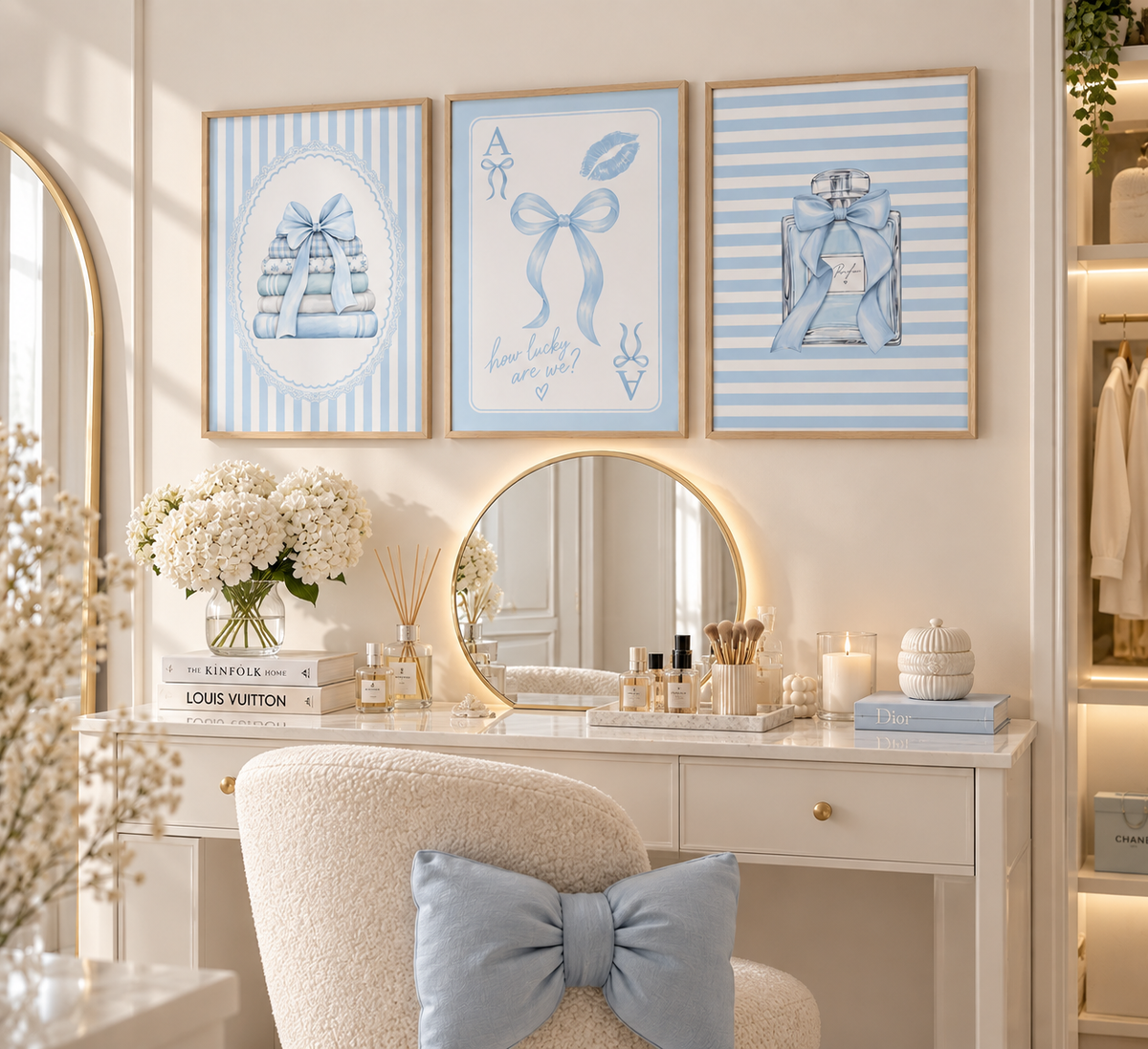 A styled vanity area with three framed blue-and-white art prints above a white desk and round mirror. The prints feature ribbon-bow illustrations, and the scene includes a cream chair with a blue bow pillow, candles, books, and floral arrangements.