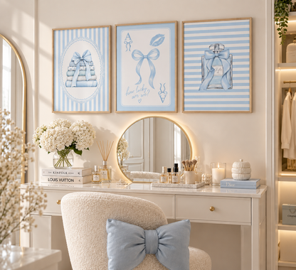 A styled vanity area with three framed blue-and-white art prints above a white desk and round mirror. The prints feature ribbon-bow illustrations, and the scene includes a cream chair with a blue bow pillow, candles, books, and floral arrangements.