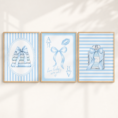 Three framed baby shower prints in a light blue and white color scheme, displayed side-by-side on a warm off-white wall with soft leaf shadows. The left print shows a stack of folded baby clothes with a bow on a striped background.