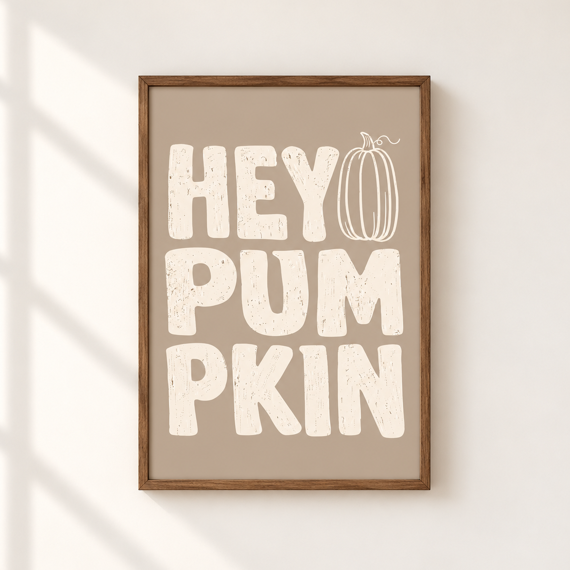Framed wall art with the phrase “HEY PUMPKIN” in large distressed cream letters on a warm taupe background, with a simple pumpkin illustration integrated into the design. The frame is wood-toned and the print is shown hanging on a light wall with soft window-shadow lighting.