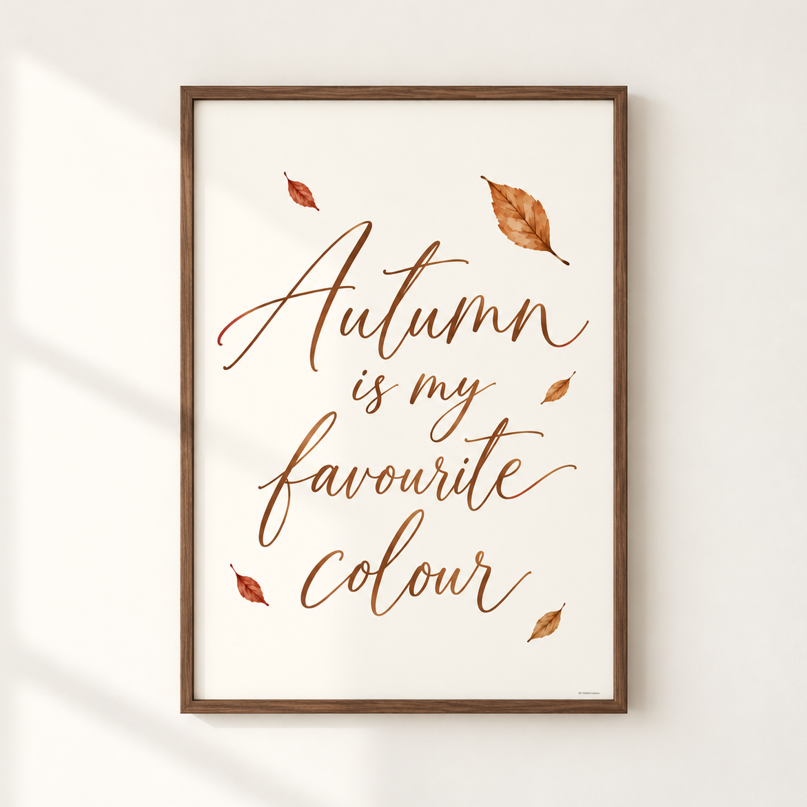 Framed wall art print with the phrase 'Autumn is my favourite colour' in copper-brown script, decorated with scattered autumn leaves, shown hanging on a light wall with soft window shadows.