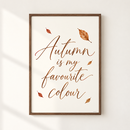 Framed wall art print with the phrase 'Autumn is my favourite colour' in copper-brown script, decorated with scattered autumn leaves, shown hanging on a light wall with soft window shadows.