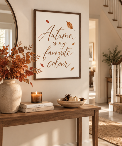 A framed autumn-themed wall art print hangs on a warm beige wall above a wooden console table. The print reads 'Autumn is my favourite colour' in elegant brown script with scattered leaf illustrations.