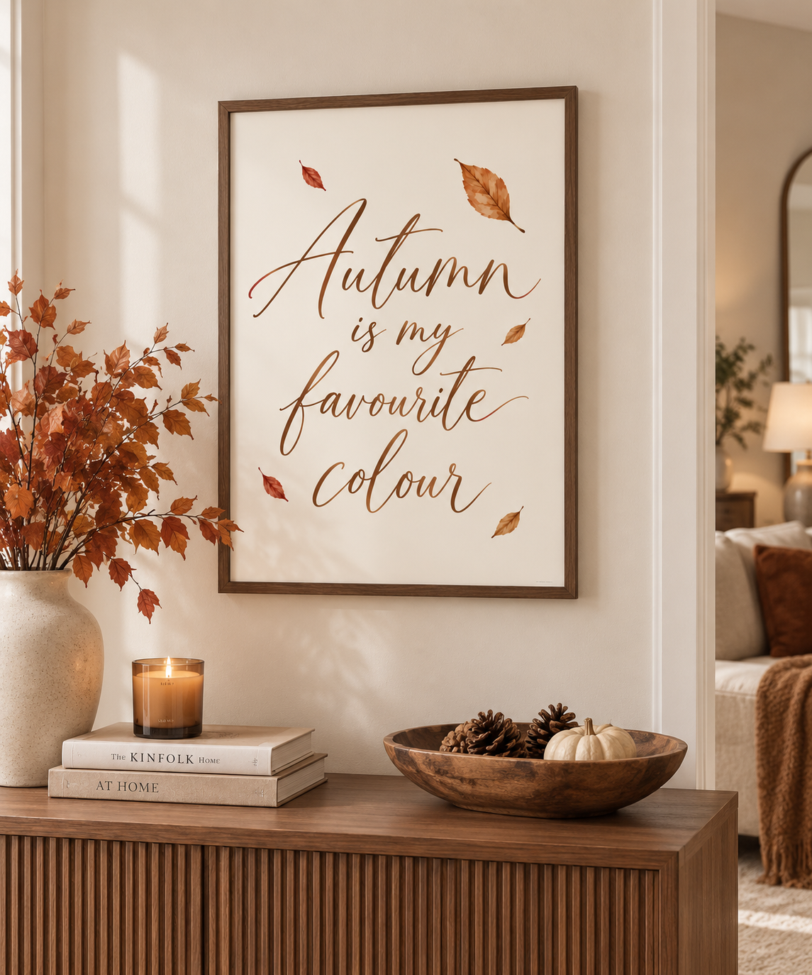 A framed autumn-themed wall art print with the phrase 'Autumn is my favourite colour' in brown script, displayed in a warm, styled living room setting above a wooden console table with fall decor.