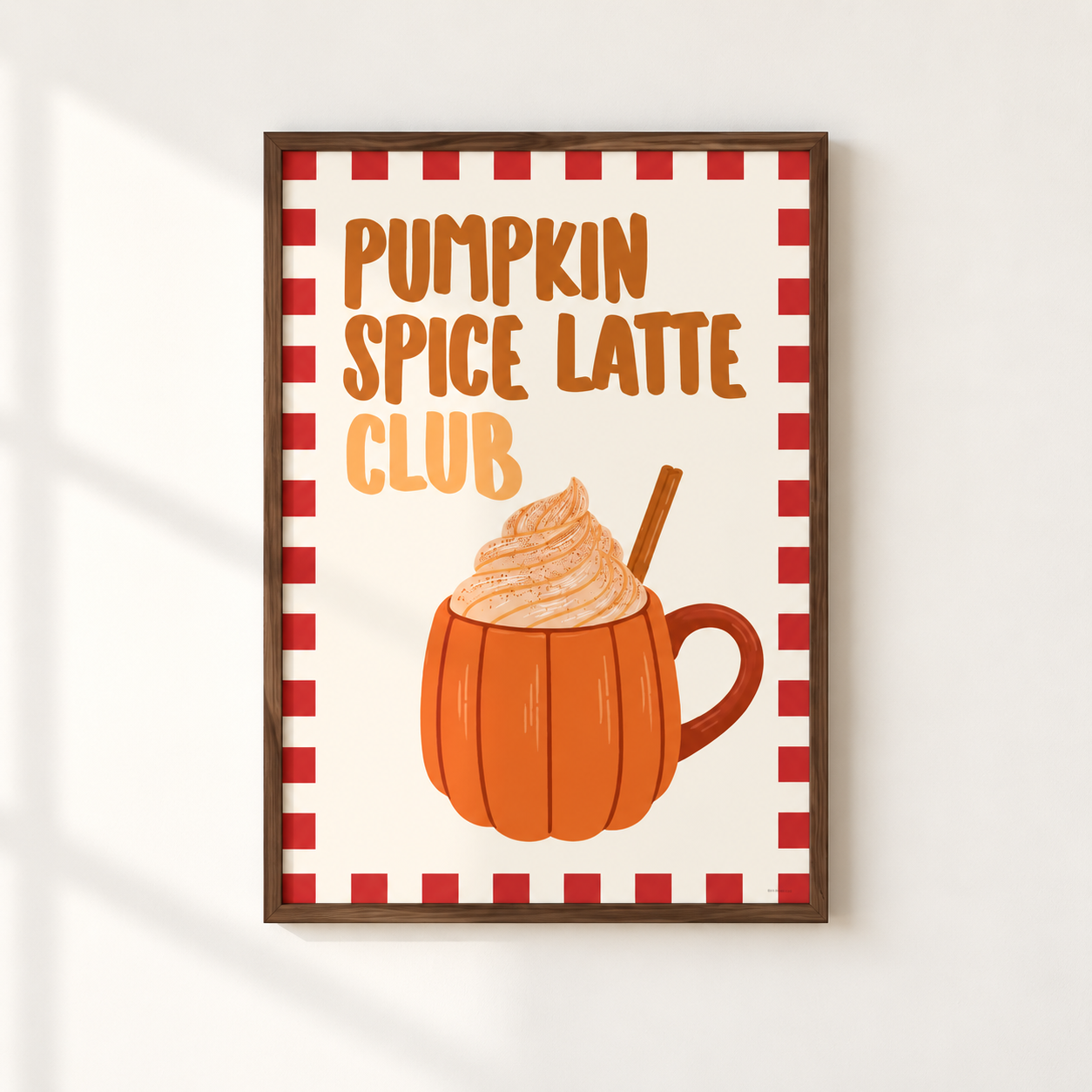 A framed autumn-themed art print on a white wall, featuring the text 'PUMPKIN SPICE LATTE CLUB' above an illustration of a pumpkin mug filled with whipped cream and a cinnamon stick.
