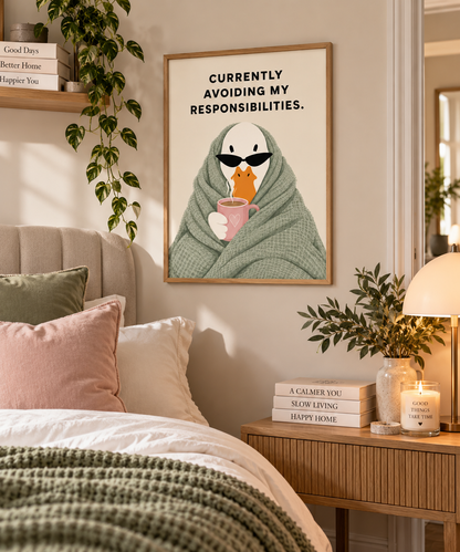 A framed wall art print featuring a cartoon bird wrapped in a green blanket and holding a pink mug, with the text 'CURRENTLY AVOIDING MY RESPONSIBILITIES.' The print is displayed in a cozy bedroom setting above a bed with pink and white pillows, a wooden nightstand, a lamp, and books.