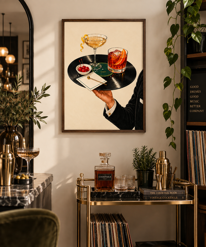 A framed art print hanging on a beige wall in a stylishly decorated bar or lounge setting. The print shows a vintage-style illustration of a waiter's arm holding a black vinyl record tray with a cocktail glass, an orange drink, a small bowl of red fruit, and a napkin.