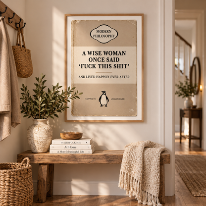 A framed vintage-style humorous poster hangs on a beige wall above a rustic wooden bench in a styled home entryway. The poster reads, 'A WISE WOMAN ONCE SAID 'FUCK THIS SHIT' AND LIVED HAPPILY EVER AFTER' with a small penguin illustration.