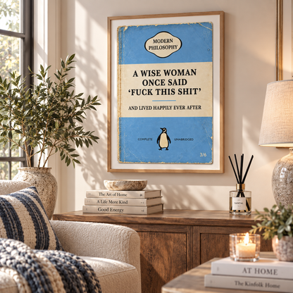 A framed art print featuring a vintage-style book cover design with the text 'A WISE WOMAN ONCE SAID FUCK THIS SHIT AND LIVED HAPPILY EVER AFTER' and a small penguin illustration, displayed on a console table in a styled living room setting.