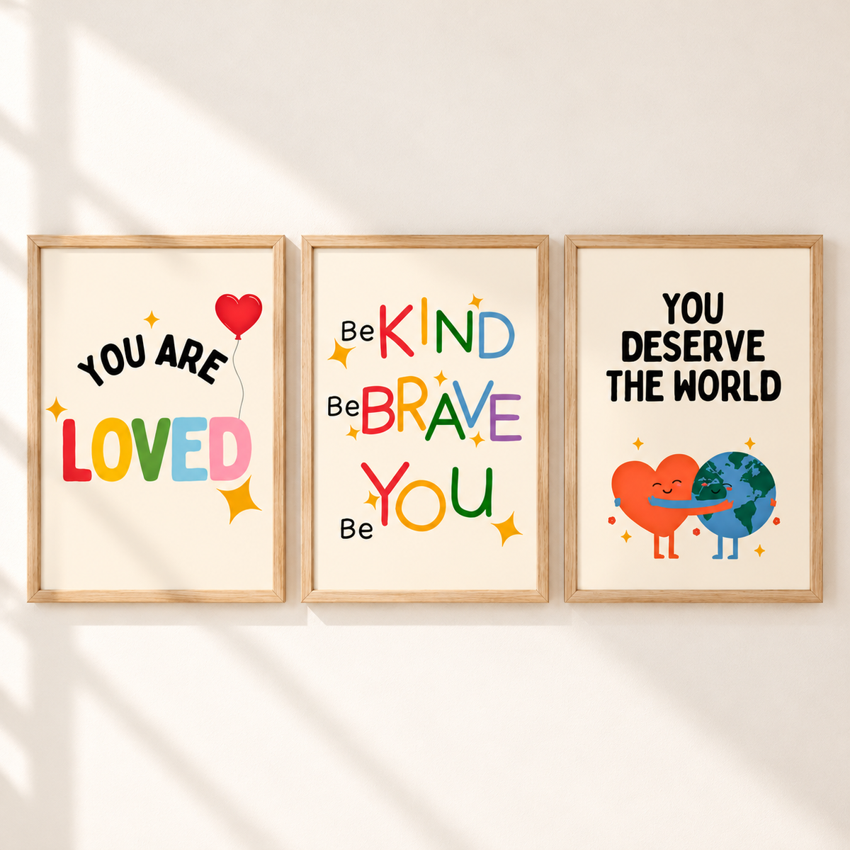 Three framed motivational wall art prints hang side by side on a light wall with soft diagonal shadows. The left print says 'YOU ARE LOVED' with a red heart balloon and multicolor lettering.