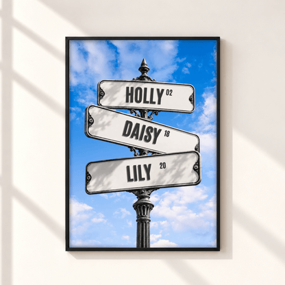 A framed wall art print showing three street signs reading HOLLY, DAISY, and LILY against a blue sky with clouds, displayed on a light wall with soft window shadows.