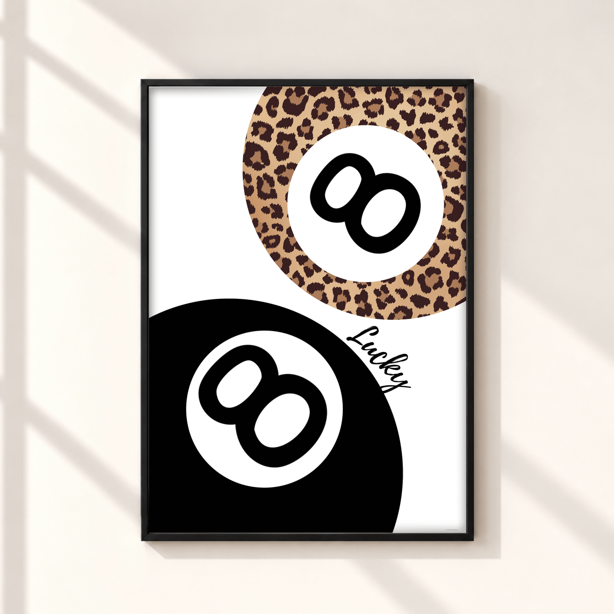 Framed wall art featuring a stylized 8-ball design with a black lower half, a leopard-print upper half, and the word 'Lucky' in script, shown hanging on a light wall with soft window shadows.