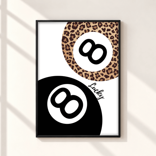 Framed wall art featuring a stylized 8-ball design with a black lower half, a leopard-print upper half, and the word 'Lucky' in script, shown hanging on a light wall with soft window shadows.