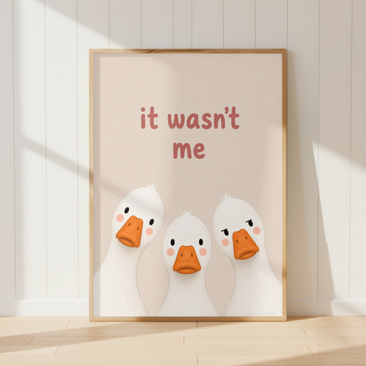 A framed art print featuring three cute white cartoon ducks with orange beaks and the text 'it wasn't me' in red, leaning against a white paneled wall on a light wood floor.