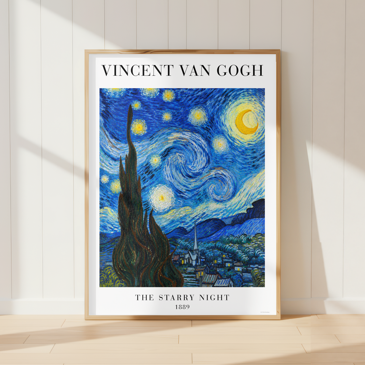 A framed art print of Vincent van Gogh's 'The Starry Night' leaning against a white paneled wall on a light wood floor, with soft sunlight casting shadows.