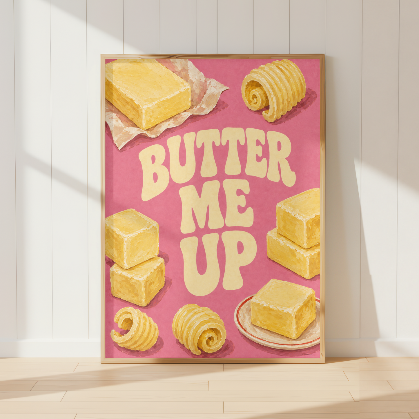 A framed retro-style wall art print leaning against a white paneled wall on a light wood floor. The print features yellow butter illustrations and the phrase “BUTTER ME UP” on a pink background.