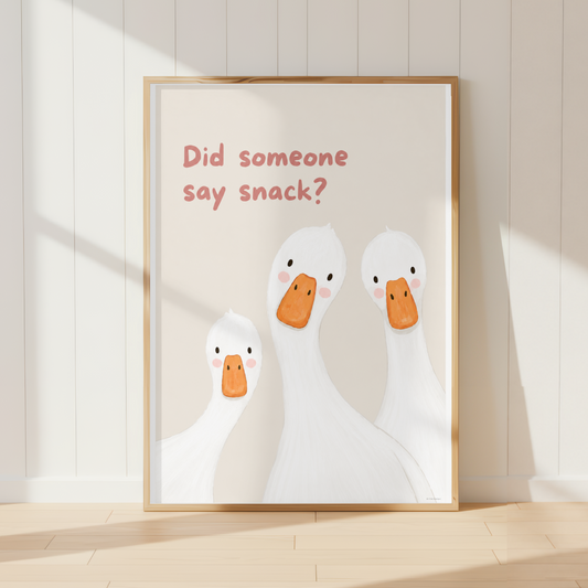 A framed kitchen art print leaning against a white paneled wall on a light wood floor. The print shows three white cartoon ducks with orange beaks and the text 'Did someone say snack?' in dusty pink.