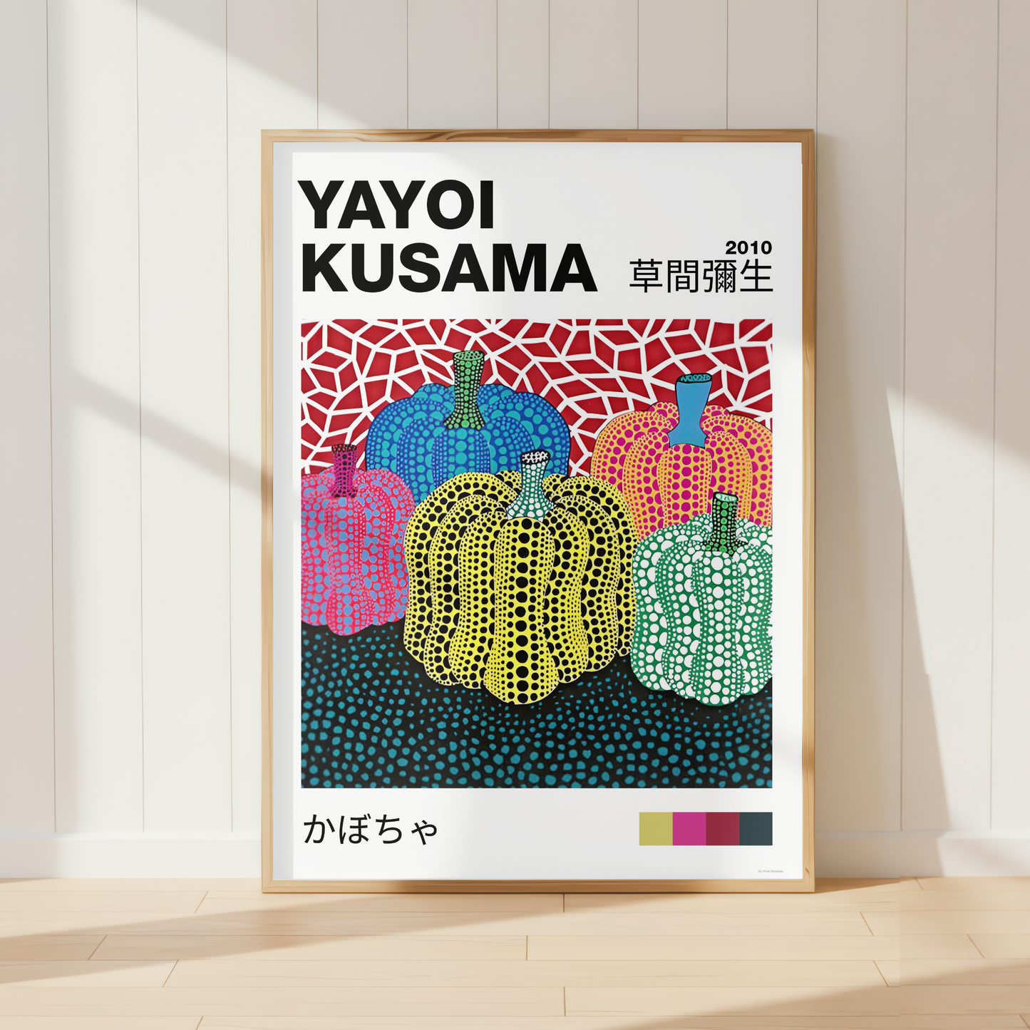 A framed art print of Yayoi Kusama's 2019 'Kabocha' (pumpkin) series, leaning against a white paneled wall on a light wood floor. The print features Kusama's signature dotted style with colorful pumpkins in pink, yellow, green, and blue, enclosed in a light wood frame.