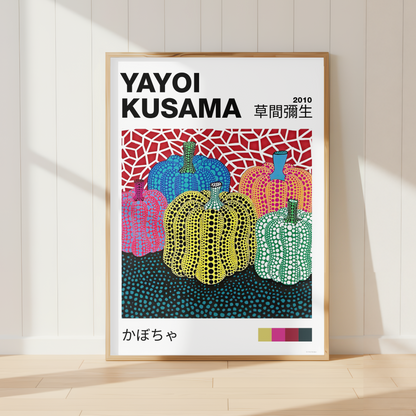 A framed art print of Yayoi Kusama's 2019 'Kabocha' (pumpkin) series, leaning against a white paneled wall on a light wood floor. The print features Kusama's signature dotted style with colorful pumpkins in pink, yellow, green, and blue, enclosed in a light wood frame.