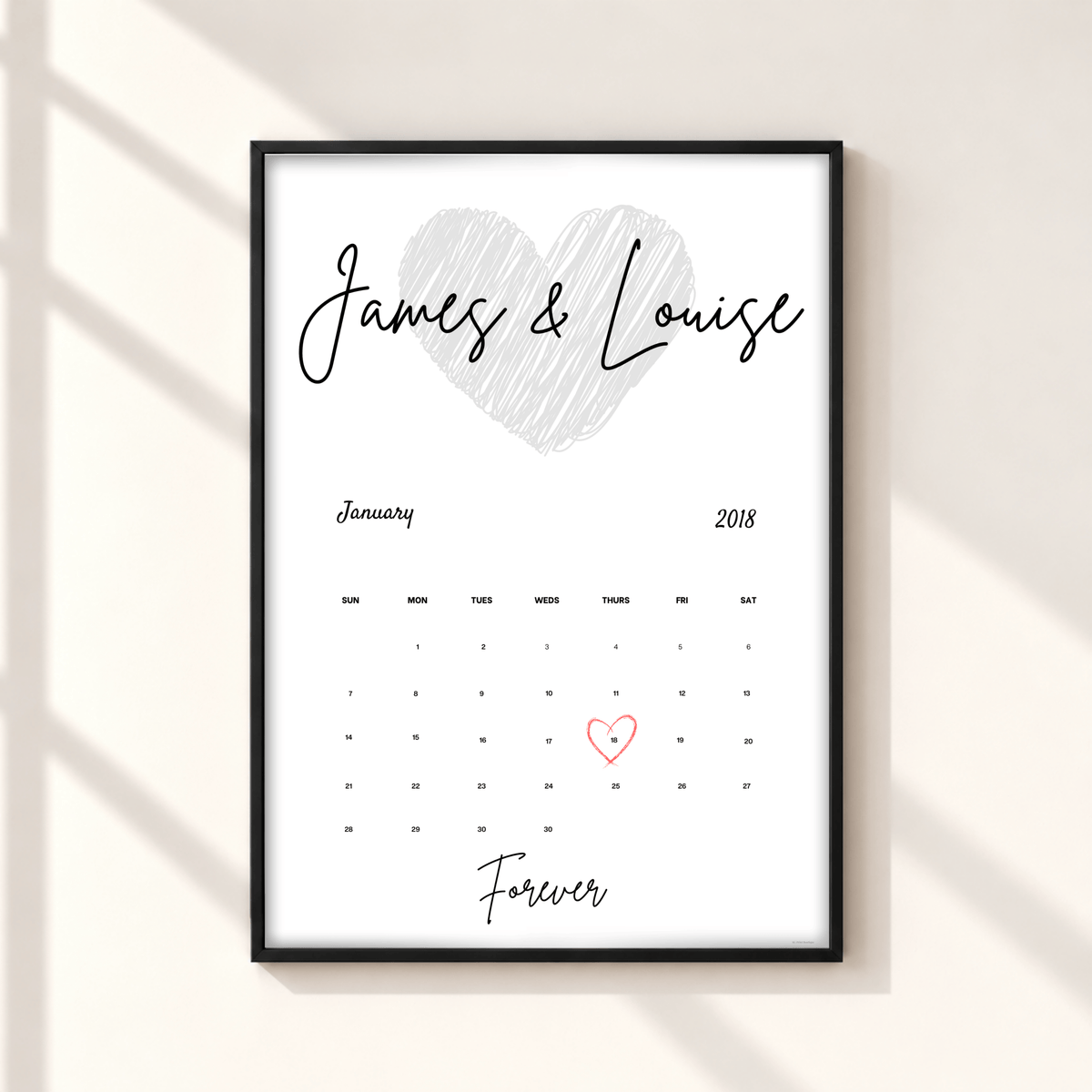A personalized framed calendar print on a light beige wall with soft diagonal shadows. The print features a white background, a black frame, a hand-drawn gray heart, the names 'James & Louise' in black script, a January 2018 calendar grid, and 'Forever' written at the bottom, with the number 1 highlighted in red.