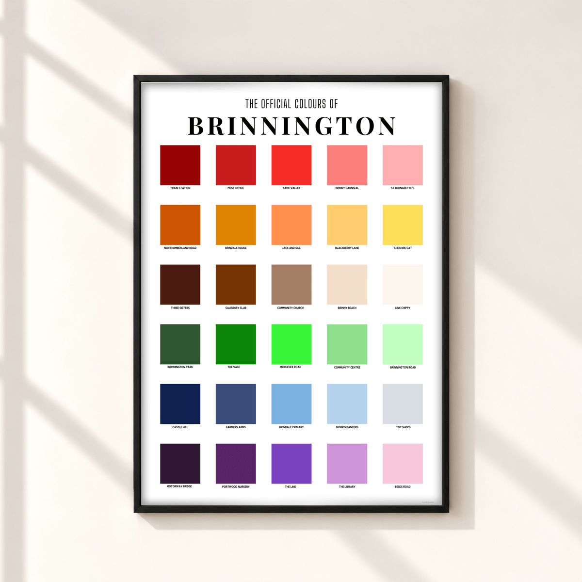 A framed print titled 'The Official Colours of Brinnington' hangs on a light beige wall. The poster features a grid of color swatches in reds, oranges, yellows, greens, blues, purples, and pinks, with printed color names beneath each square.