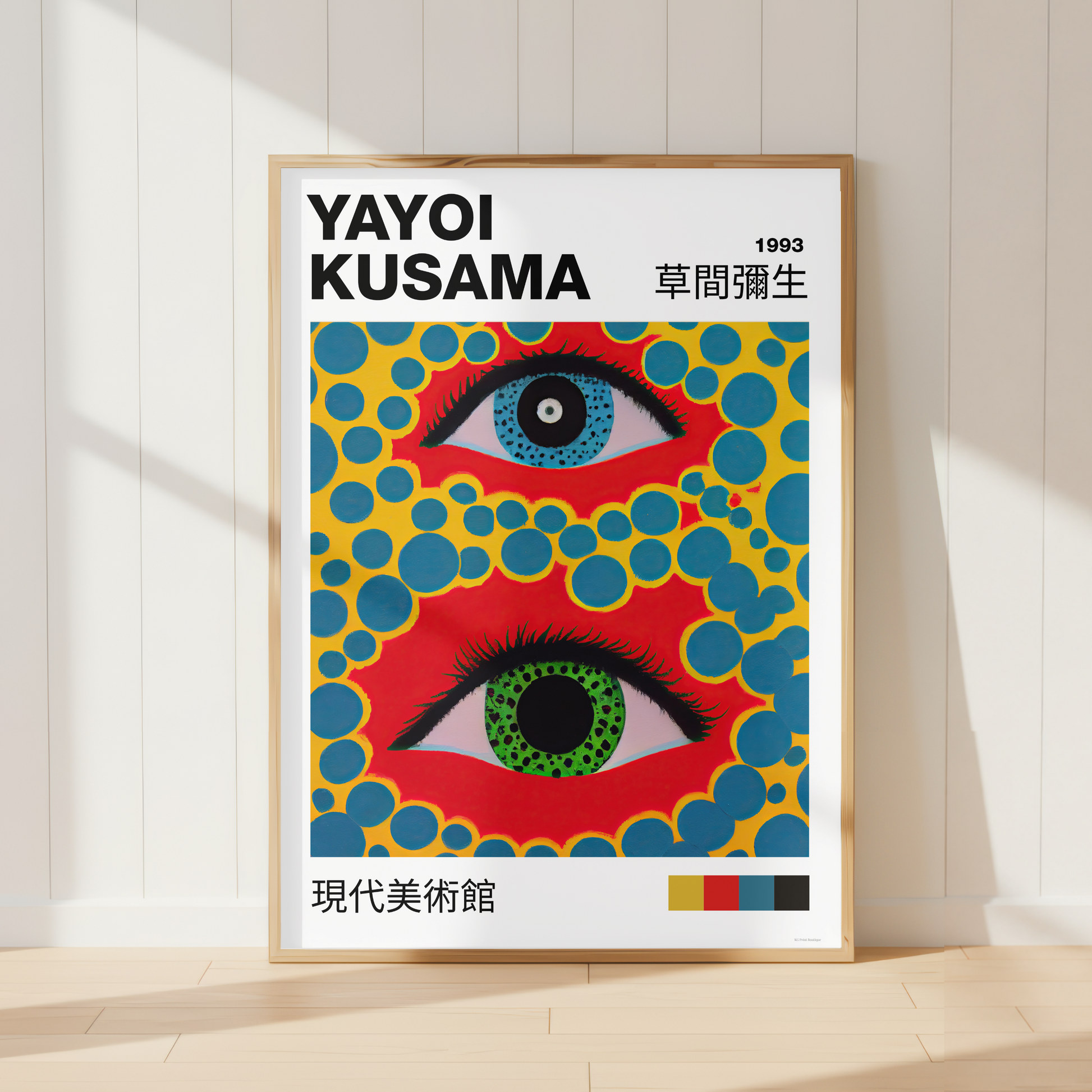A framed art print of Yayoi Kusama's 1993 exhibition poster, leaning against a white paneled wall on a light wood floor. The poster features two stylized eyes with red, blue, green, and yellow patterns.
